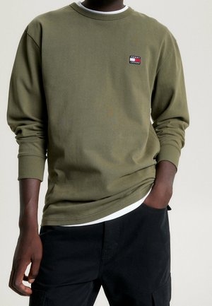 Mężczyzna ubrany w oliwkową, długorękawową bluzę Tommy Jeans z małą naszywką z logo na piersi, czarne spodnie i białą koszulę wystającą spod dołu bluzy.