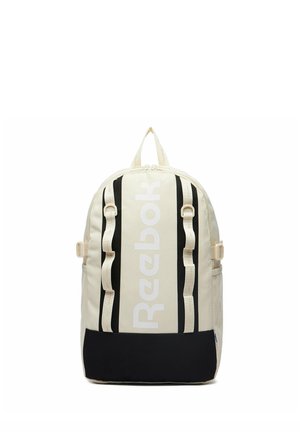 Beigefarbener und schwarzer Reebok-Rucksack mit vertikalem weißen Logo, verstellbaren Riemen, seitlichen Netzfächern und oberem Tragegriff vor weißem Hintergrund.