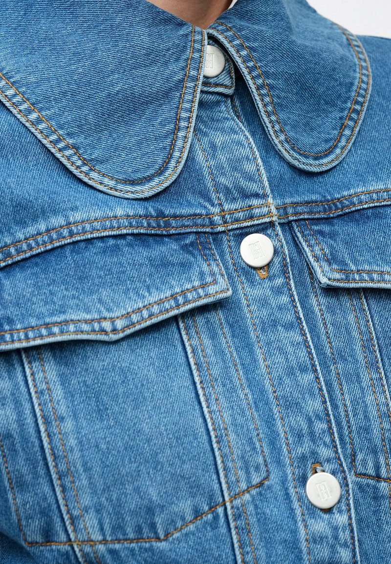 Denim jacket featuring a large, rounded collar, light blue hue, white buttons, and contrasting stitching around pockets and seams.