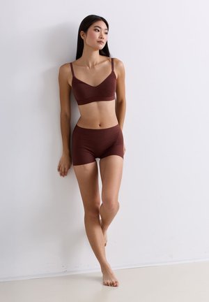 Frau in braunem Sport-BH und Shorts steht barfuß, lehnt sich an eine weiße Wand und blickt entspannt zur Seite.