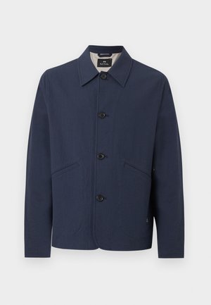 Mand iført marineblå jakke over blå skjorte med beige vidde bukser og hvide sneakers, stående med den ene hånd i lommen.