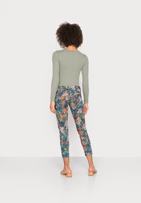 Kvinde med krøllet hår iført en ribbet grøn bluse med lange ærmer, blomster-mønstrede cropped bukser og beige hælede sandaler, stående med ryggen til.