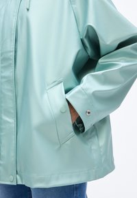 Πράσινο φρέσκο μπουφάν βροχής με λεία, γυαλιστερή επιφάνεια, που διαθέτει τσέπη με κουμπί κλικ και μανίκια με κουμπιά, και άνετη εφαρμογή με ρυθμιζόμενο κουκούλα.