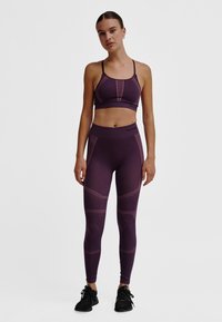 Paarse sportbeha en legging met contrasterende roze stiksels, gestructureerde details en een nauwsluitend ontwerp. Draagt zwarte sportschoenen.