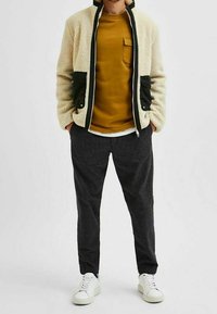 Fleecová bunda v krémové barvě se zelenými akcenty a kapsami, nošená přes hořčicovou košili. Šedé kalhoty s vzorem a bílé tenisky doplňují outfit.