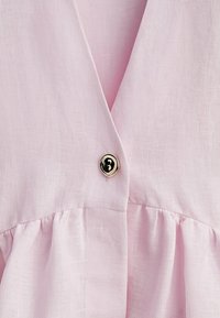 Robe rose clair avec un décolleté en V, des manches courtes à volants et un détail de bouton doré brillant à la taille. Tissu doux et texturé.