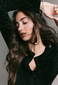 Top nero con texture e scollo con bottoni, caratterizzato da un motivo sottile. Capelli lunghi che fluiscono, con un braccialetto d'argento al polso.