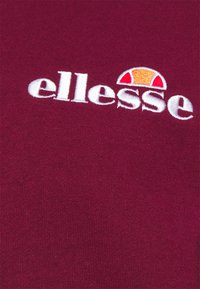 Bordeaux-Stoff mit weißem, gesticktem "ellesse"-Logo und halbkreisförmigem Design in Orange und Rot über dem Text.