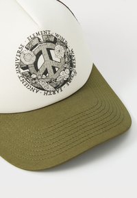 Gorra con una parte superior crema y visera en verde oliva, que presenta un símbolo de paz circular rodeado de diseños florales y texto en negro.