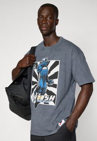 Pelēks pārāk liels t-krekls ar zilu supervaroņa grafiku uz melnbalta fona, sarkanas Nike zīmes akcentiem un mīkstu kokvilnas tekstūru.
