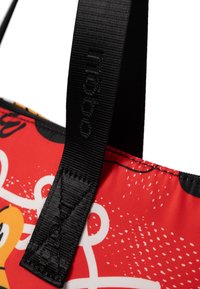 Rood gestippelde tote bag met een zwarte strap en een gegraveerd logo. Het patroon heeft abstracte vormen en witte accenten op een glanzend oppervlak.