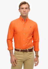 Mężczyzna w jasnopomarańczowej koszuli z guzikami z podwiniętymi rękawami, beżowych spodniach, plecionym brązowym pasku i zegarku na ręku, stojący na białym tle.