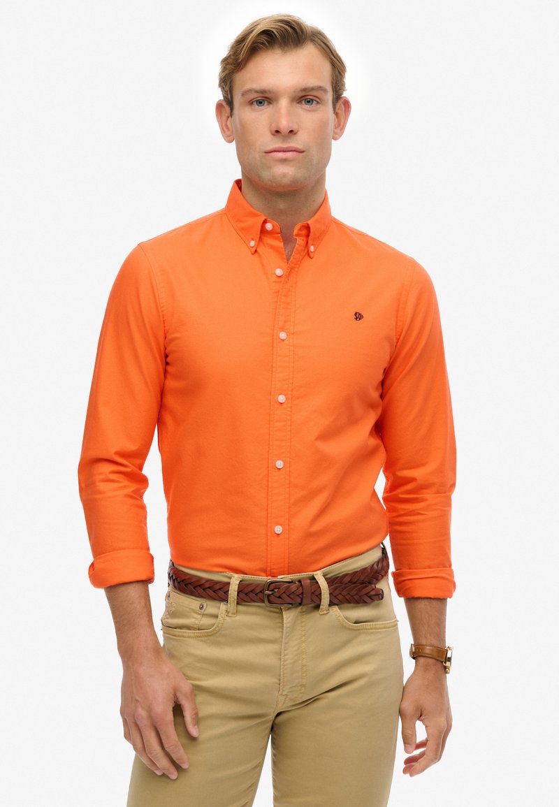 Mężczyzna w jasnopomarańczowej koszuli z guzikami z podwiniętymi rękawami, beżowych spodniach, plecionym brązowym pasku i zegarku na ręku, stojący na białym tle.