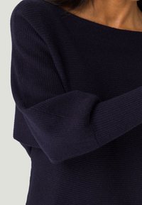 Maglioncino blu navy con scollo ampio, texture a coste e vestibilità comoda. Le maniche sono leggermente arrotolate e raccolte sui polsini.