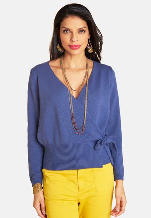 Frau trägt einen blauen Wickelpullover, der seitlich gebunden ist, gelbe Hosen, mehrlagige Perlenketten, bunte Ohrringe und ein breites goldenes Armband.