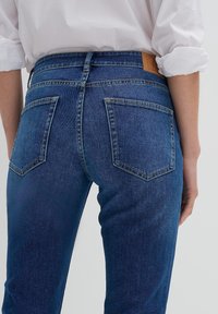 Bakifrån av en person som bär blå denimjeans och en vit skjorta med uppvikta ärmar mot en neutral bakgrund.