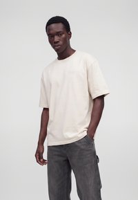 Giovane uomo che indossa una t-shirt oversize beige chiaro a maniche corte e jeans larghi grigio scuro, in piedi davanti a uno sfondo chiaro e uniforme.