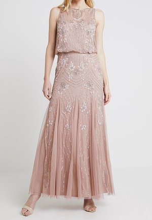 Žena v bezrukávových, dlouhých růžových šatech s květinovými a geometrickými flitrovými zdobeními, doplněná otevřenými lodičkami v tělové barvě.