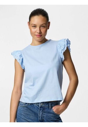 Jeune femme portant un haut sans manches bleu clair avec manches brodées à volants et un jean bleu, posant avec une main dans la poche.