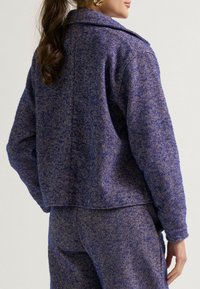 Wollmischung-Jacke im lila Fischgrätenmuster mit Kragen und abgesenkten Schultern; kombiniert mit passenden Hosen, die eine strukturierte Oberfläche zeigen.
