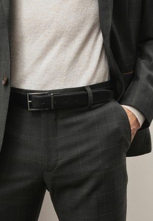 Mørkternet jakkesæt med en struktureret overflade, parret med en beige sweater og et sort læderbælte med en sølvspænde.