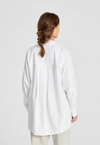 Camicia bianca a maniche lunghe con collo alto, vestibilità ampia e dettaglio a pieghe sul retro; realizzata in tessuto leggero e liscio.