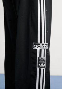 Melna jaka ar baltu Adidas trīs svītru dizainu, spiedpogām un logotipa plāksteriem. Gluda auduma, brīva piegriezuma.