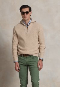 Man in beige kwart-rits trui, blauwe gestreepte shirt, groene broek, bruine riem, polshorloge en donkere zonnebril, staand tegen een effen achtergrond.