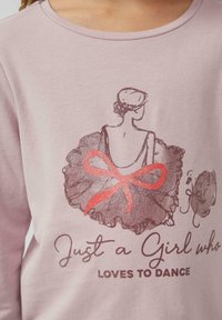 Różowa koszula z długim rękawem, z grafiką tancerki baletowej w tutu z falbankami i błyszczącą czerwoną kokardą. Tekst "Just a Girl who Loves to Dance" w ciemnym druku.