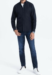 Tmavě modrá košile na knoflíky s kapsou na hrudi, v kombinaci se středně modrými džínami a bílými teniskami s barevně kontrastními detaily.