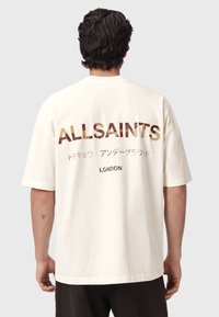 Μπεζ βαμβακερό t-shirt με μεγάλο κείμενο "ALLSAINTS" σε σκούρο καφέ και χρυσό στην πλάτη, με επιπλέον κείμενο στα ιαπωνικά και τη λέξη "LONDON" από κάτω.