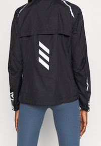 Musta kevyt takki, jossa on tuuletusaukko selässä, heijastavat valkoiset yksityiskohdat ja vinoraitoja. Ominaisuuksiin kuuluvat pitkät hihat ja sileä pinta.