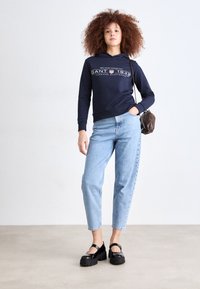 Hanorac bleumarin cu text grafic alb, blugi deschisi cu talie inalta, si pantofi cu platforma negri. Are par ondulat si o geanta maro crossbody.