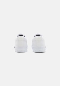 White sneakers featuring a smooth leather upper, circular eyelets, and a textured heel tab, with branding on the sole. Minimalist design.