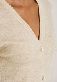 Nahaufnahme eines cremefarbenen Strickcardigans mit V-Ausschnitt und zwei runden Knöpfen auf der Brust und Schulter einer Person.