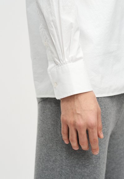 Δεξί χέρι χαλαρό στο πλευρό, φορώντας ένα λευκό πουκάμισο με μακριά μανίκια και κουμπωμένο μανσέτι και γκρι παντελόνι.