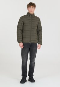 Doudoune vert olive avec des coutures horizontales, un col montant et des poches zippées, associée à un jean gris foncé et des chaussures noires.