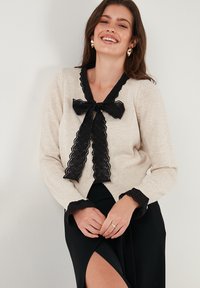 Beige knitted cardigan with black lace trim, bow at the neck, and scalloped cuffs, worn over a black skirt. Smooth texture and fitted design.