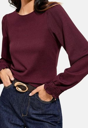 Femme portant un haut bordeaux à manches longues avec des poignets smockés, rentré dans un jean bleu foncé avec une ceinture noire dotée d'une grande boucle dorée.