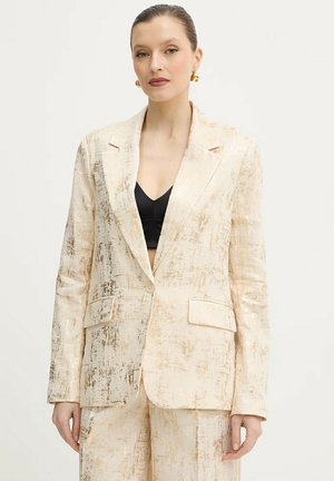 Donna che indossa un blazer crema e oro dalla texture strutturata e pantaloni abbinati, sopra un crop top nero, con orecchini a cerchio dorati.