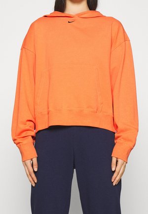 Femme portant un sweat à capuche orange à manches longues avec poche avant et petit logo noir, associé à un pantalon bleu marine foncé.