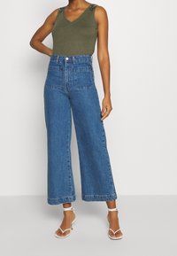 Camiseta sin mangas verde oliva con escote en V y detalles decorativos en los hombros, combinada con unos pantalones vaqueros de mezclilla azul de pierna ancha y cintura alta, y tacones blancos con tiras.