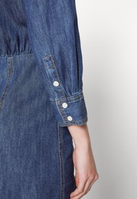 Rukáv džínové bundy se sladěným manžetem třemi bílými knoflíky, kontrastním šitím a uvolněným střihem z texturované látky.