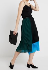 Svart camisole med spetskant som kombineras med en plisserad kjol i svart, grön och blå. Tillbehör inkluderar en svart clutch och svarta klackar.