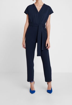 Kobieta ubrana w granatowy kombinezon z głębokim dekoltem w kształcie V, krótkimi rękawami, paskiem w talii i jasnoniebieskimi czółenkami na spiczastym obcasie, stojąca na białej podłodze.