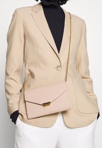 Béžové sako s kabelkou na zlatém řetízku. Kabelka je světle růžová s plochým designem, klopovým zavíráním a zlatými kovovými prvky.