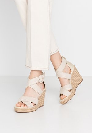 Fødder iført beige fletvævede kilehælssandaler med ankelremme og spænder, kombineret med off-white korte bukser mod en ensfarvet baggrund.