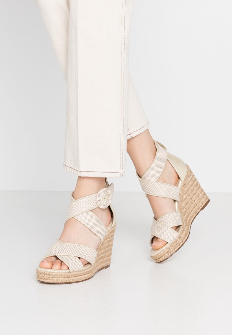 Pieds chaussés de sandales compensées en tissu beige avec brides à la cheville et boucles, associés à un pantalon court blanc cassé sur un fond uni.