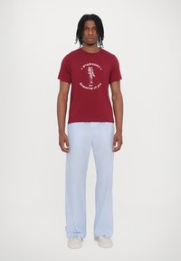 Burgundy cotton t-shirt with white graphic print, paired with blue and white striped wide-leg pants. Model stands against a plain background.