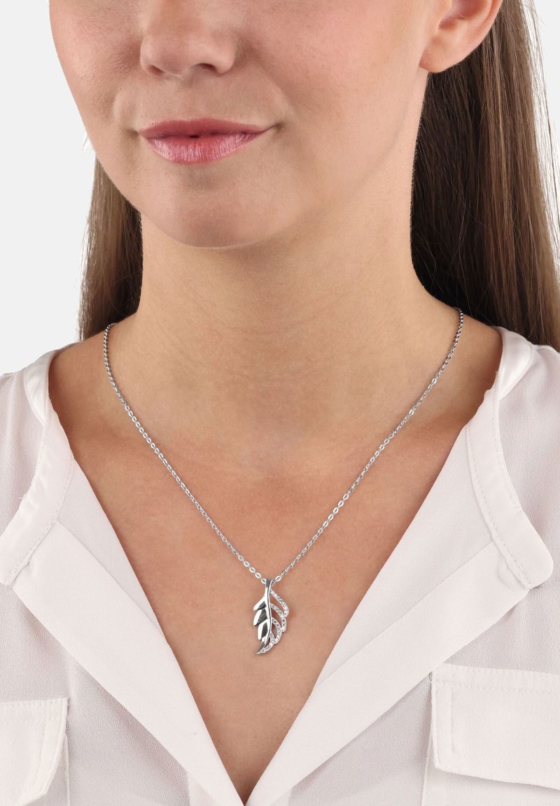 Halsband med hängande i form av silverlöv prytt med små kristaller, bärs av en kvinna i en vit blus med v-ringning.